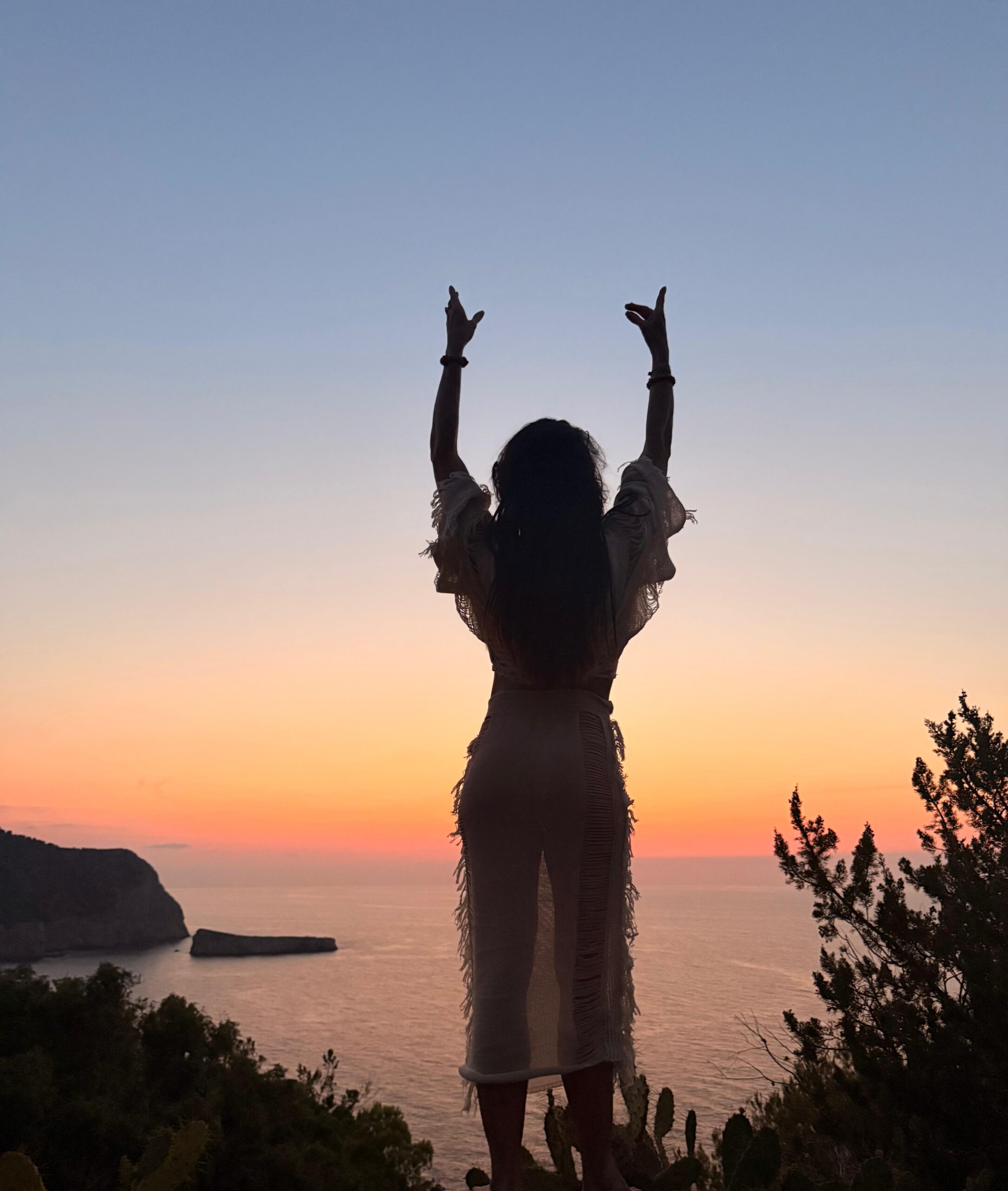Woman standing on a cliff edge at sunset, arms raised to the sky in a moment of presence and release
